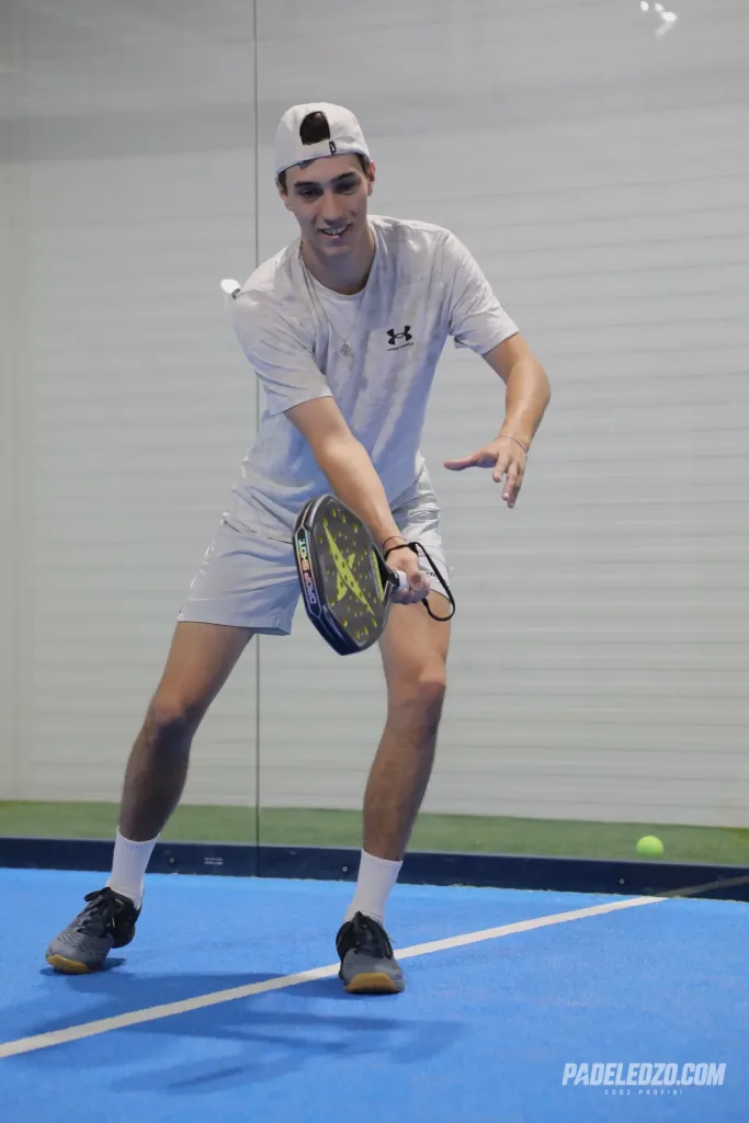 Erik fehér sapkában és világos sportruhában, ütés közben a fedett padelpályán